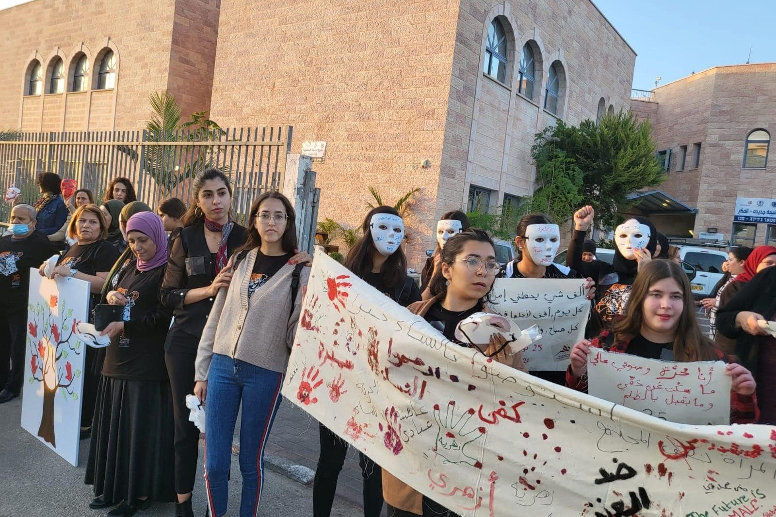 Eine Gruppe von Frauen und Mädchen steht in einer Reihe vor einem großen Gebäude mit Backsteinfassade. Einige tragen weiße Masken, die symbolisch für Stille oder Unsichtbarkeit stehen könnten, während andere normale Kleidung tragen. Einige der Teilnehmerinnen halten Schilder mit arabischen Schriftzügen und Symbolen, die auf Gewalt oder Frieden hinweisen. Die Frau vorne links trägt einen lilafarbenen Hijab und hält ein Bild mit einem Baum und roten Handabdrücken. Die Atmosphäre wirkt ruhig, aber entschlossen, da die Frauen offensichtlich für eine friedliche Sache eintreten.
