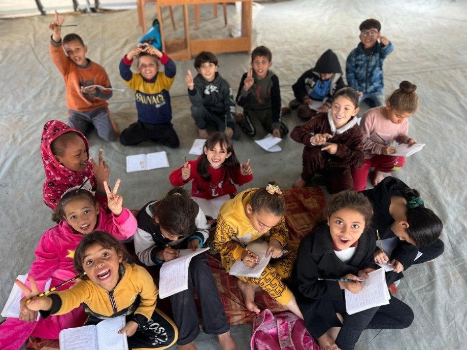 Kinder sitzen auf dem Boden eines Raums, einige zeigen das Victory-Zeichen, andere schreiben in Hefte, Umgebung schlicht mit Stuhl und Tisch im Hintergrund.