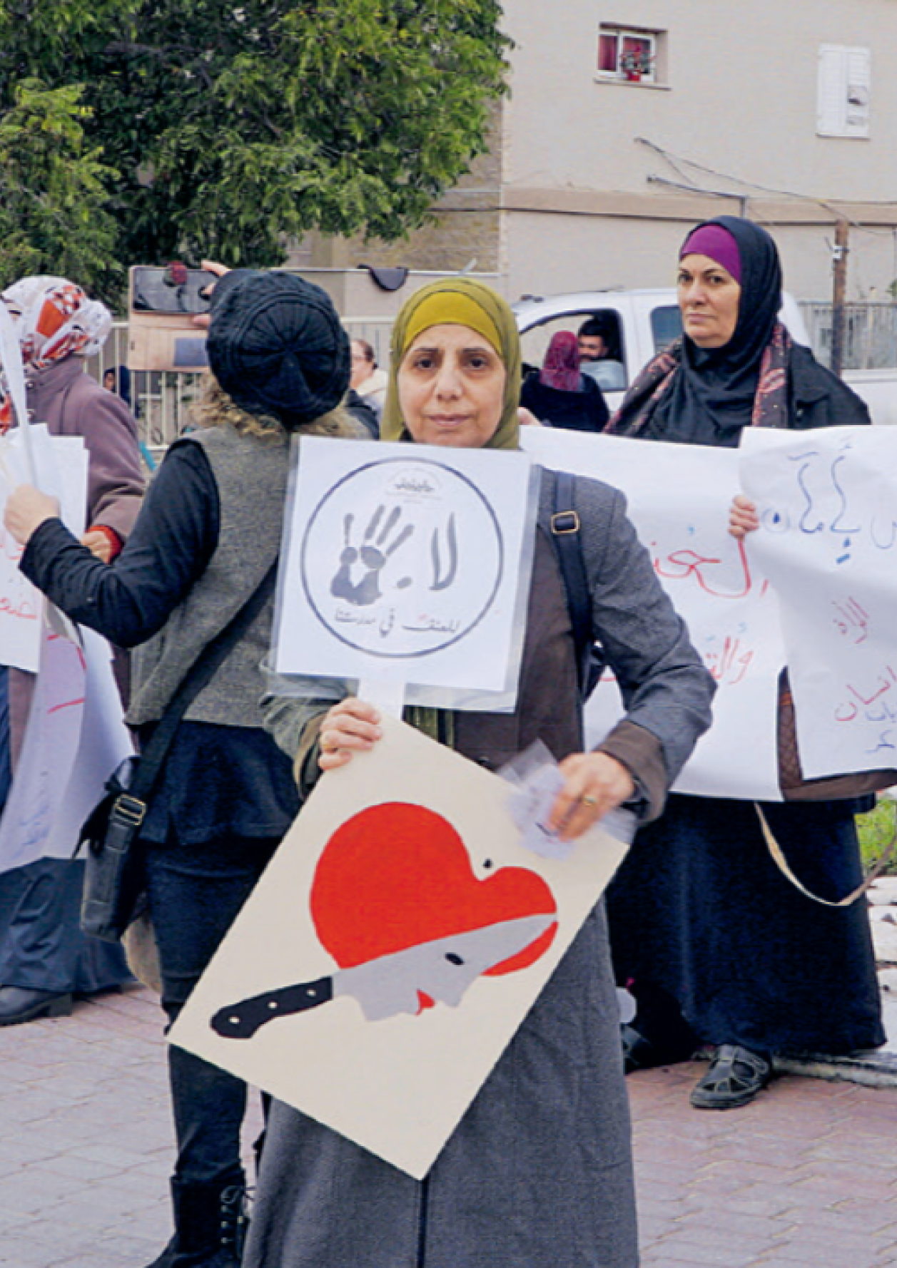 Eine Gruppe von Frauen hält Protestplakate auf einer Strasse hoch. Die Plakate enthalten arabische Schriftzüge, Zeitungsartikel und symbolische Bilder, darunter ein Herz mit einem Messer. Die Demonstration scheint sich gegen Gewalt, insbesondere gegen Frauen, zu richten. Im Hintergrund sind Autos, Gebäude und Bäume zu sehen.